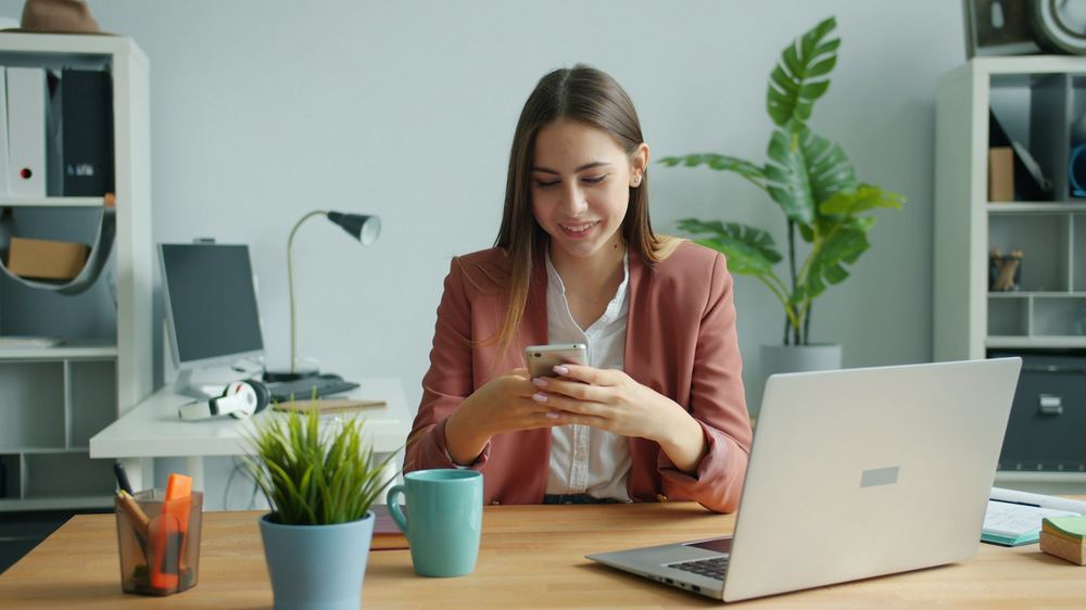 Uśmiechnięta kobieta sprawdza konto w telefonie przy biurku